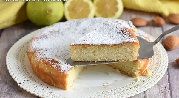 Torta delle monache soffice e profumata, cosparsa di zucchero a velo, con limoni e mandorle sullo sfondo.