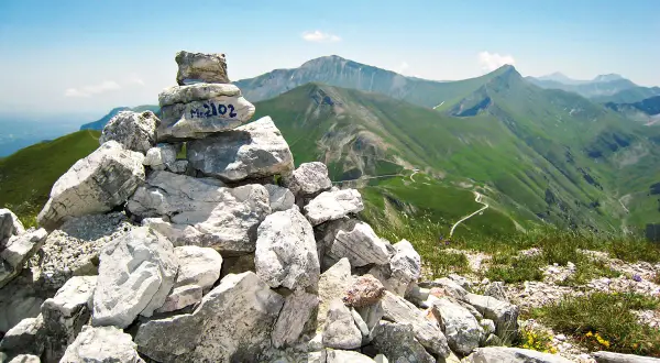 Cairn con M. 2102, simbolo di un'avventura di trekking nei Monti Sibillini. Panoramica mozzafiato sulle verdi valli e sentieri tortuosi.