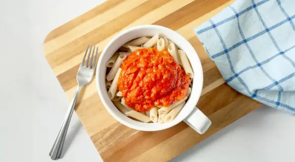 Makaron penne z sosem pomidorowym w białej misce, obok widelec i ściereczka.