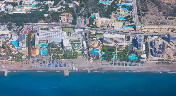 Vy från ovan över ett stort vattenland och hotellkomplex vid Rhodos kust. Solstolar och parasoller kantar stranden.