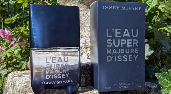 Flakon i pudełko perfum Issey Miyake L'Eau Super Majeure d'Issey na kamiennym postumencie, otoczone zielenią i różowymi kwiatami.