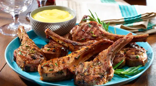 Chuletillas de cordero a la parrilla, jugosas y doradas, servidas en un plato azul con romero fresco y una salsa amarilla.