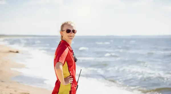 Livräddare i rött och gult står på stranden, redo för sol och bad. Vågorna skvalpar vid hennes fötter.