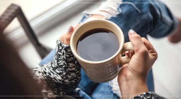 En person med slitna jeans och en stickad tröja håller en mugg med kaffe på morgonen.