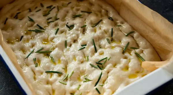 Luftiger focaccia teig mit Rosmarin und Olivenöl in einer Backform, bereit zum Backen.