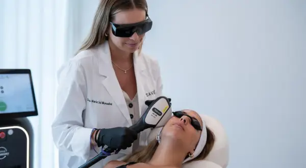 Médica estética con gafas protectoras realiza tratamiento facial a paciente con dispositivo láser.