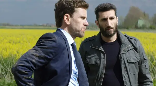 Två män står i en gul rapsåker. En av dem bär kostym, den andra en militärjacka. Scen från avdelning q filmer.