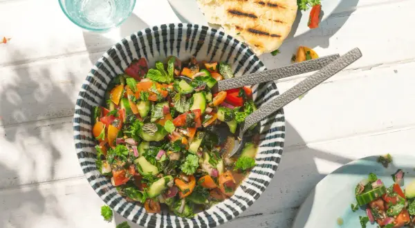 Frischer Tomaten Gurken Salat mit Kräutern, serviert in einer Schüssel mit blau-weißen Streifen. Daneben ein Glas Wasser und gegrilltes Fladenbrot.