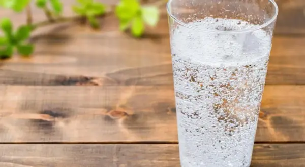 Un verre rempli d'eau gazeuse pétillante, sur une table en bois. Une branche avec des feuilles vertes floues est visible à gauche.