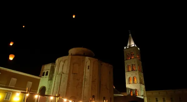 Nocne niebo nad Zadarem rozświetlają lampiony. Widać zabytkowe budowle, w tym okrągły kościół i wieżę.