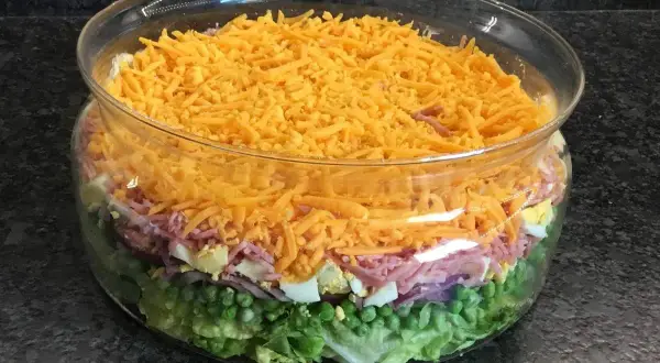 Une magnifique salade 7 étages dans un saladier en verre, avec des couches de laitue, petits pois, oignons rouges, jambon, œufs durs et fromage râpé.