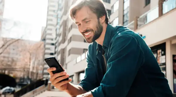 Uśmiechnięty mężczyzna z zarostem trzyma w dłoni czarny telefon, przeglądając coś z zaciekawieniem. W tle nowoczesne budynki.