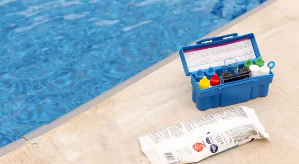 Pool test kit and shock treatment next to a blue pool. Learn how to shock a pool for clear, clean water.