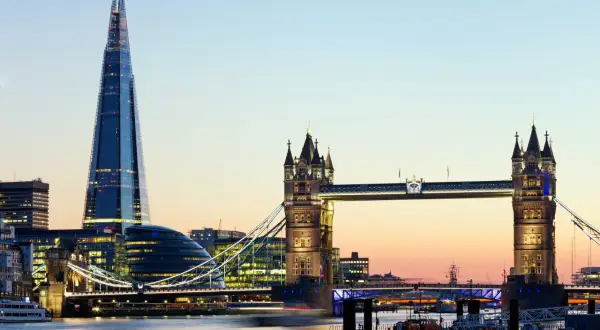 Widok na Tower Bridge i The Shard w Londynie o zmierzchu, z rzeką Tamizą i łodziami w tle.