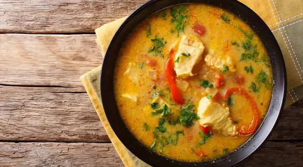 Bol de blanquette de poisson crémeuse avec des morceaux de poisson, des poivrons rouges et du persil, servi sur une serviette jaune sur une table en bois.