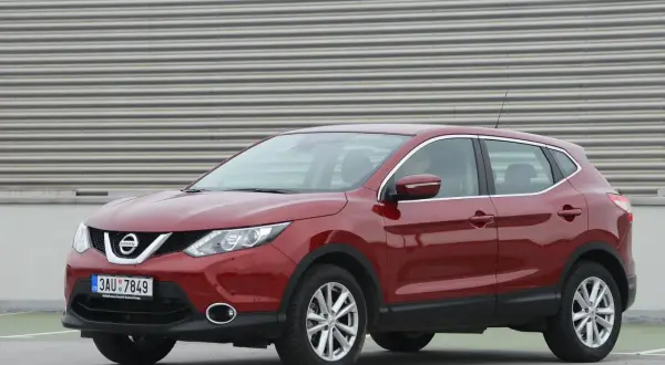 Czerwony Nissan Qashqai J11, z widocznym wykazem żarówek w przednich reflektorach, zaparkowany na tle falistej ściany.