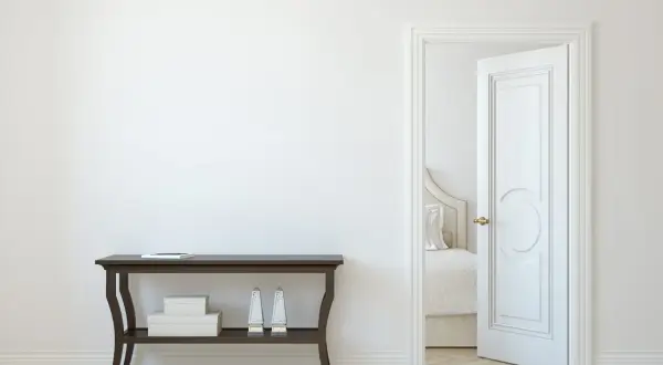 Un corridoio con un tavolino scuro e una porta aperta che rivela una stanza da letto. La porta interna è dipinta di bianco, con un design classico.