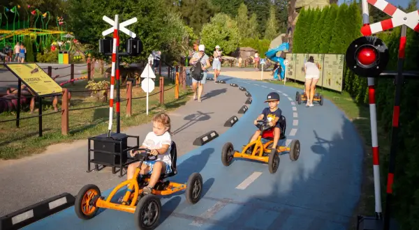 Dzieci ścigają się gokartami na torze w parku rozrywki. W tle widać inne atrakcje i spacerujących ludzi.