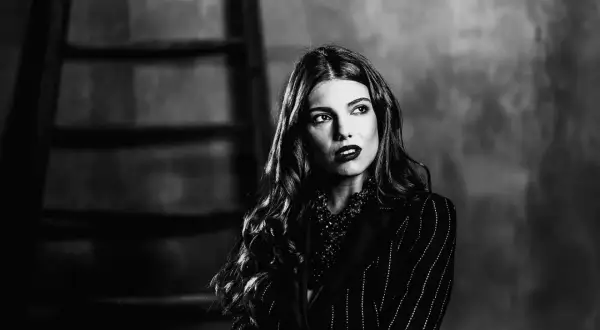 Ritratto bianco e nero di una donna con capelli mossi e labbra scure, indossa una giacca a righe e una collana vistosa.