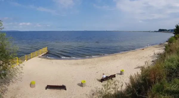 Plaża dla psów z łagodnym zejściem do morza. Na piasku ławki i kosze. W oddali widać falochron i statki.