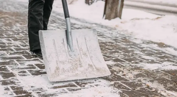 Osoba w czarnych spodniach odśnieża chodnik łopatą.