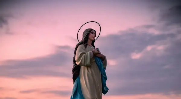 Figurka Matki Boskiej z aureolą na tle różowego nieba. W dłoniach trzyma błękitną szatę, jakby w modlitwie "Anioł Pański".