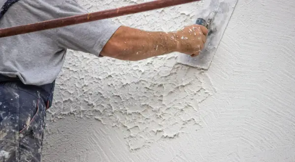 Artigiano applica intonaco esterno con cazzuola, creando texture rustica. Diversi tipi di finitura intonaco esterno sono visibili.