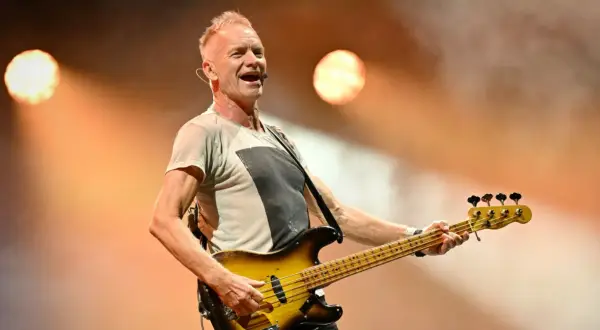Sting, lider zespołu The Police, gra na gitarze basowej na scenie, oświetlony ciepłym światłem.