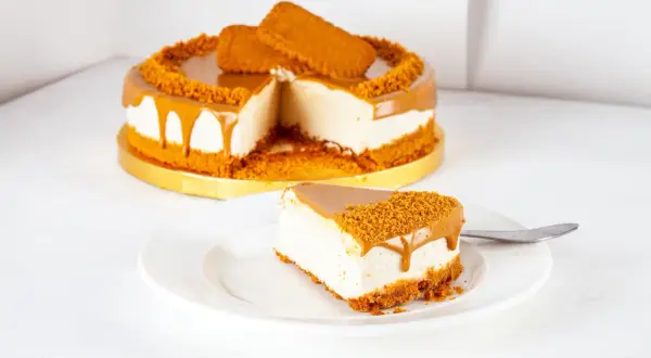 Ein Stück cremiger Lotus Cheesecake mit Karamell und Keksbröseln auf einem Teller, daneben der ganze Kuchen.
