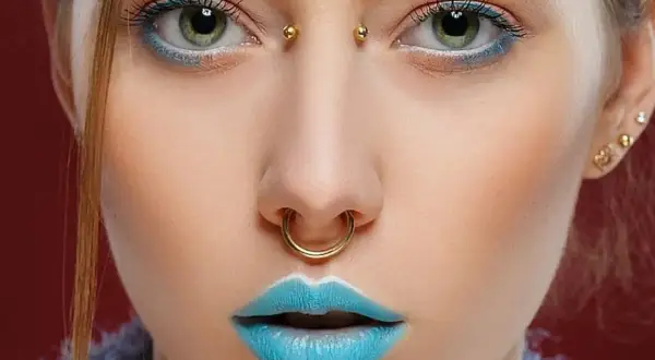 Dziewczyna z zielonymi oczami, błękitnymi ustami i złotymi kolczykami: septum, bridge piercing i w uchu.