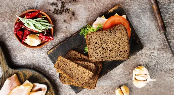 Pyszny chleb z razowej mąki pszennej, idealny na kanapki. Obok składniki: pomidor, ser, wędlina, czosnek, papryczki chili i zioła.