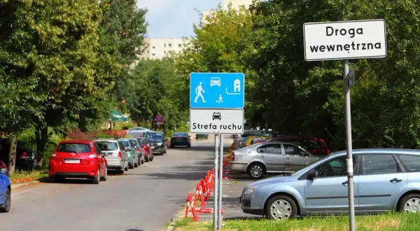 Droga wewnętrzna z oznaczeniem strefa zamieszkania. Samochody parkują wzdłuż ulicy, otoczonej drzewami.