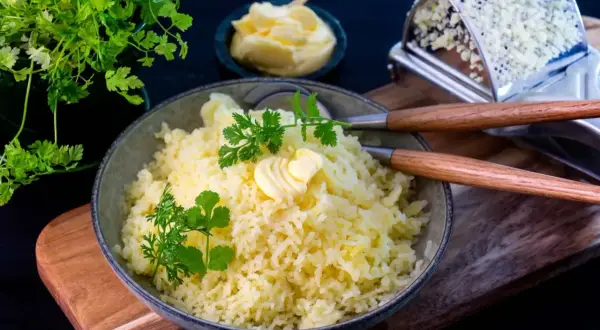 En skål med fluffig pressad potatis, toppad med en smörklutt och färsk persilja. Bredvid syns en smörskål och en rivare.