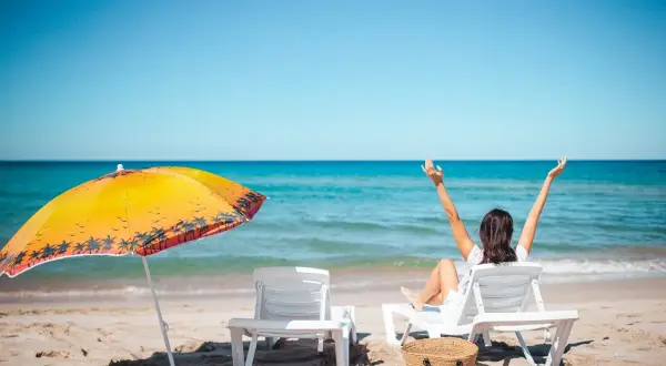 Kobieta cieszy się słońcem na plaży, zastanawiając się, ile dni urlopu jeszcze jej zostało. Obok leżaki i parasol.