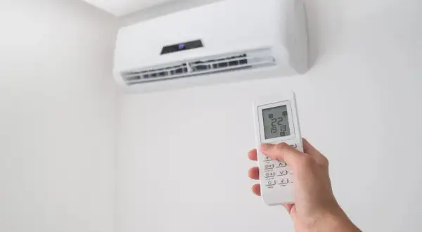 Dłoń trzymająca pilot do klimatyzacji na tle białej ściany z jednostką klimatyzacyjną.