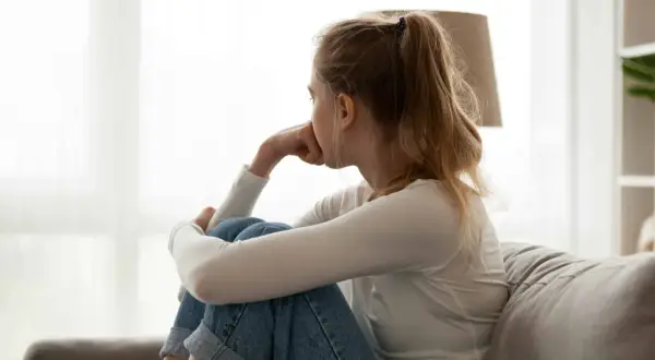 Meisje zit op de bank, met de knieën opgetrokken en de kin op de handen, een teken van overprikkeling symptomen.