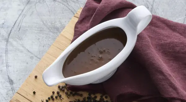 En vit såsslev fylld med mörk brunsås, bredvid en hög med svartpepparkorn på en träskärbräda. Perfekt för ditt brunsås recept.