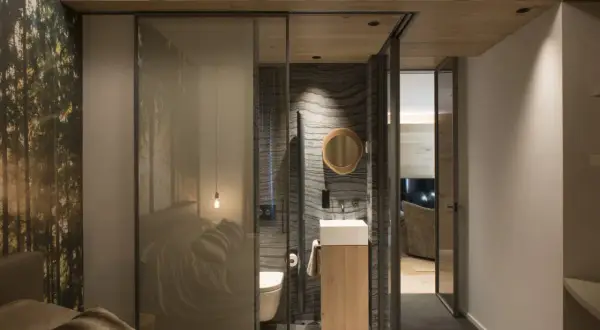 Bagno in camera di 2 mq con pareti in pietra, lavabo sospeso e specchio rotondo. La zona notte è separata da una parete in vetro con stampa foresta.