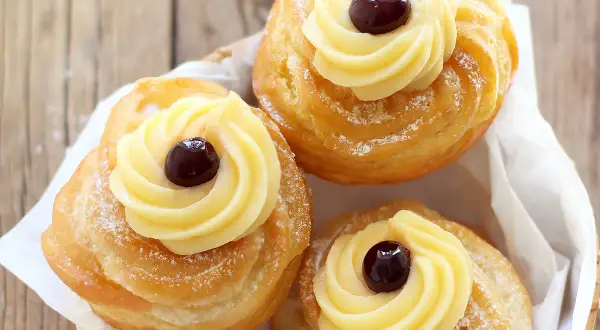Tre zeppole fritte napoletane, dorate e spolverate di zucchero a velo, con crema pasticcera e una ciliegina.