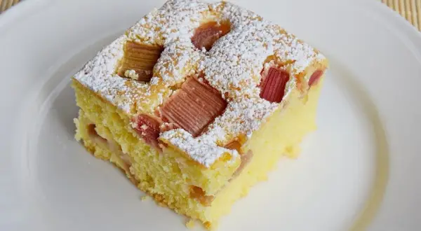 Puszyste ciasto z rabarbarem, posypane cukrem pudrem, na białym talerzu. Idealne na letnie popołudnie.