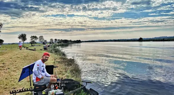 Mężczyzna w czerwonej czapce siedzi nad wodą, gotowy do łowienia ryb metodą picker feeder. W tle widać innych wędkarzy i piękne, chmurne niebo.