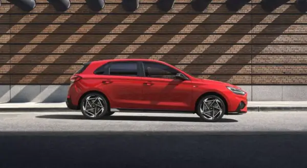 Czerwony Hyundai i30 Hatchback zaparkowany na tle ceglanej ściany z geometrycznymi cieniami.