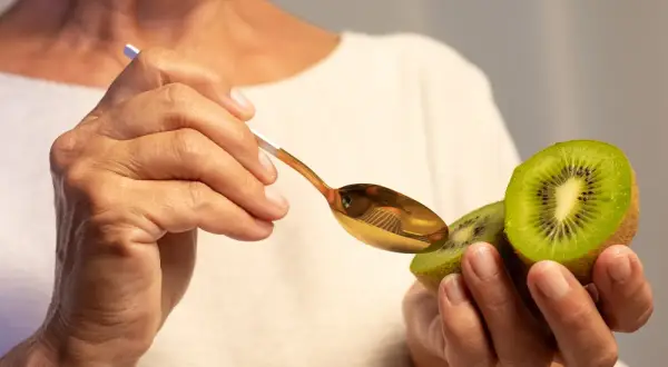 Dłonie trzymają połówkę kiwi i złotą łyżeczkę. Owoc jest bogaty w witaminy, np. C i K.