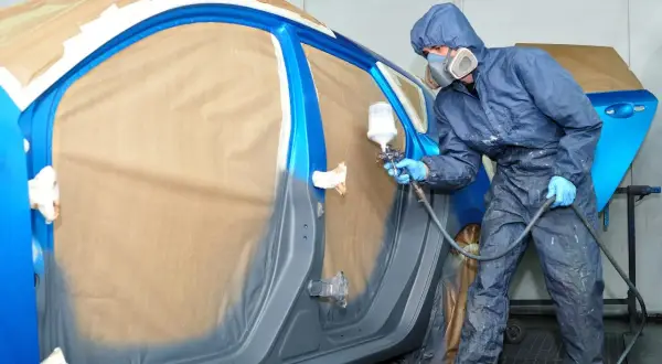 Pittore in tuta blu spruzza vernice su un'auto blu. Il costo per cambiare il colore della macchina sul libretto è un dettaglio importante.