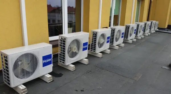 Rząd białych jednostek klimatyzacyjnych Haier na dachu, na tle żółtej ściany budynku.