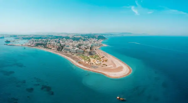 Luftfoto över Rhodos kustlinje med en sandstrand, hotell och turkost vatten. En segelbåt syns i vattnet, perfekt för Rhodos aktiviteter.