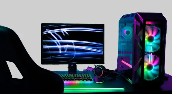 Gracz wpatrzony w monitor z niebieskimi smugami światła. Obok komputer z kolorowymi wentylatorami. Idealne miejsce na fajne słowa po angielsku na nick.
