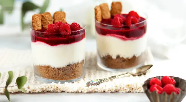 Zwei Gläser mit Schichtdessert: Keksboden, cremige Füllung, Himbeersoße und frische Himbeeren. Perfekte nachtisch ideen für jeden Anlass.