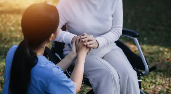 Un infermiere tiene le mani di una paziente su una sedia a rotelle, un gesto di conforto e cura. L'età non conta, l'assistenza è l'essenza.