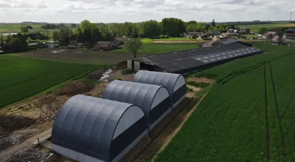 Tre tunnel stalla grigi, moderni, affiancati in un campo verde, con un grande edificio agricolo sullo sfondo.